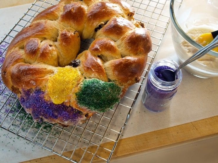 A circular King Cake is being decorated on a wire cooling rack with brightly colored sugar. Nearby sits a container of colorful sugar with a spoon resting inside.