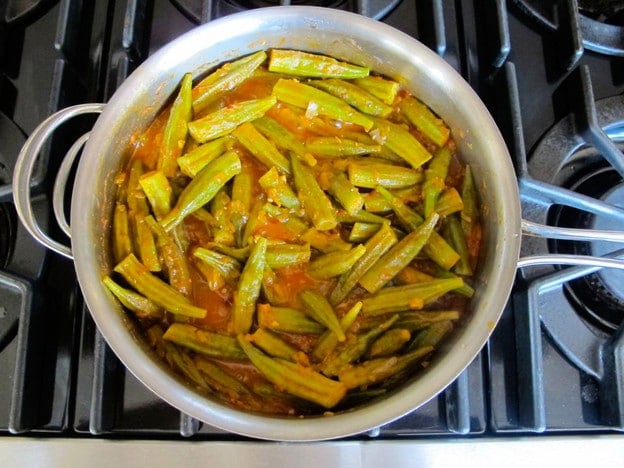 A stainless steel pot on a stove contains okra pieces cooked in a tomato-based sauce.