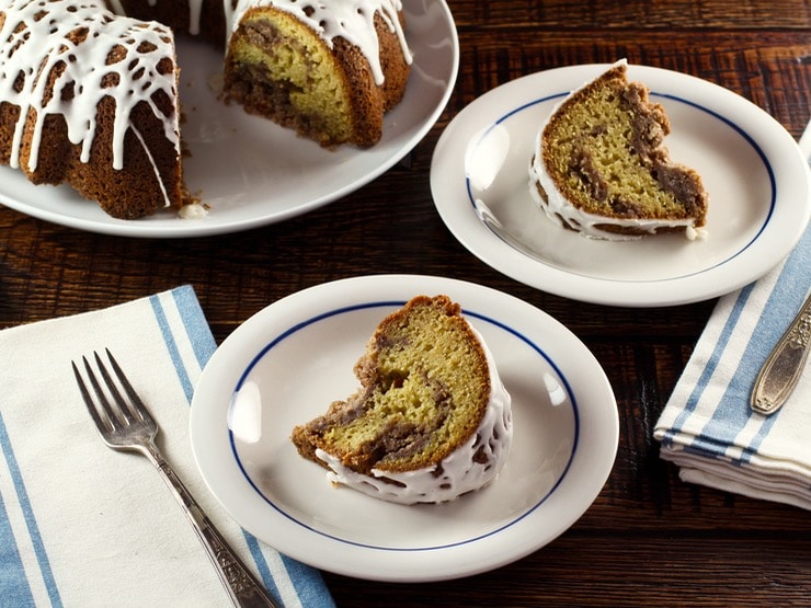 Slice of sour cream coffee bundt cake with drizzled white icing on a plate, another piece on plate in background, fork and napkin in foreground, whole cake in background.