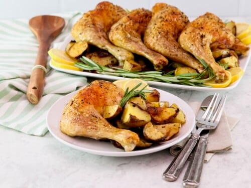Horizontal shot - in foreground, a small plate with a whole roasted chicken leg, roasted potatoes, and rosemary with, fork, knife, and linen napkin beside it. In background, a platter of roasted chicken legs on a bed of roasted potatoes, lemon, and fresh rosemary. Wooden spoon and kitchen towel beside the platter.
