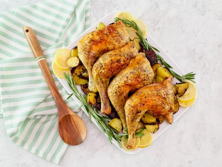 Overhead shot - platter of roasted chicken legs on a bed of roasted potatoes, lemon, and fresh rosemary. Wooden spoon and linen towel beside the platter.