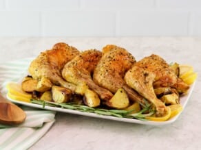 Platter of roasted chicken legs on a bed of roasted potatoes, lemon, and fresh rosemary. Wooden spoon and linen towel beside the platter.