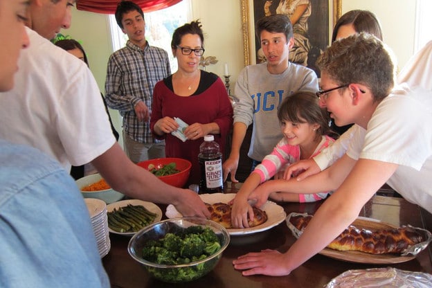 Female teacher leads a group of students in the Shabbat blessings around a dining table covered with food - including challah. Eight students gather around, and some are touching the food.