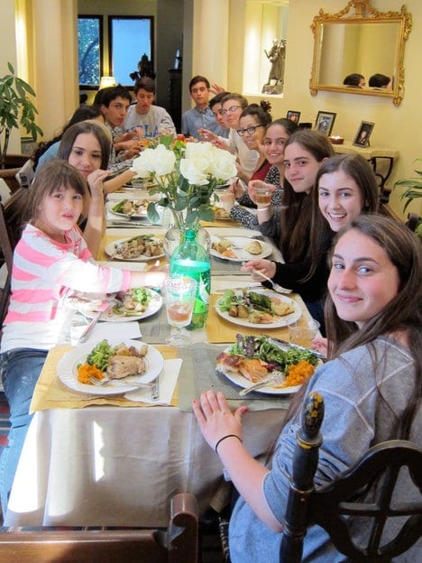 A long table with smiling students seated, enjoying a Shabbat dinner together. Plates covered with roasted rosemary chicken and side dishes.
