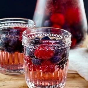 Two decorative glasses of Vanilla Berry Sparking Sangria on a wooden cutting board with wooden spoon, pitcher in background.