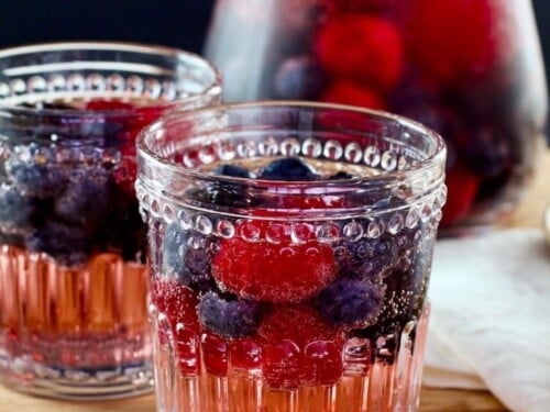 Two decorative glasses of Vanilla Berry Sparking Sangria on a wooden cutting board with wooden spoon, pitcher in background.