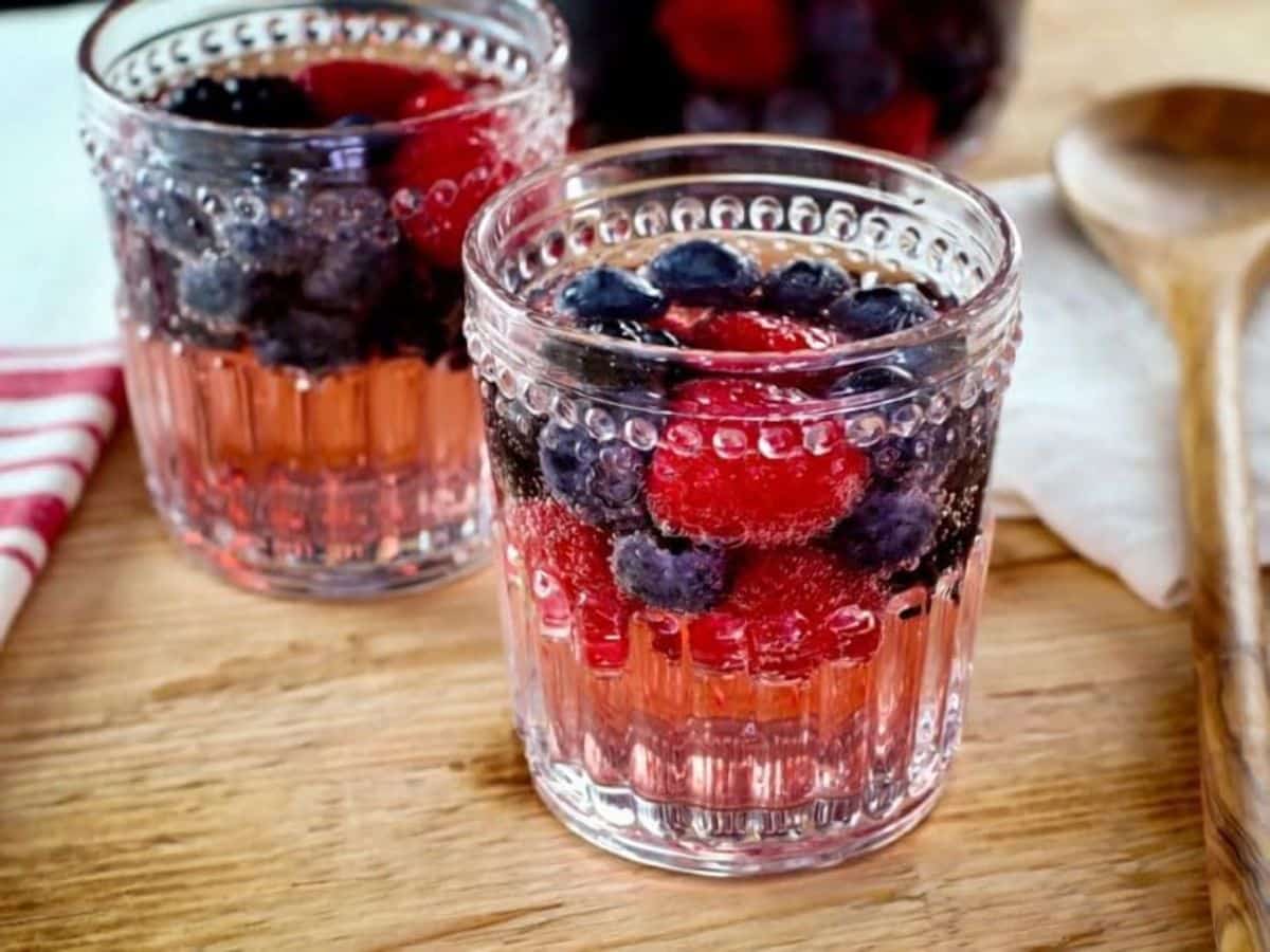 Two decorative glasses of Vanilla Berry Sparking Sangria on a wooden cutting board with wooden spoon, pitcher in background.