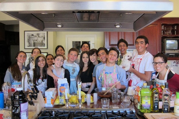 Group of 13 high school students gathered with Tori in her kitchen to prepare Shabbat dinner, smiling for the camera with their teacher. One student holds a small dog.