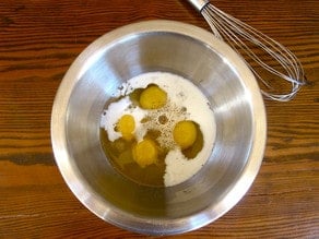 Four cracked eggs in a large mixing bowl together with milk or half and half - on a wooden table. Whisk lays beside the bowl.