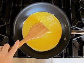 Hand with wooden spatula pushing cooked egg to the center of a nonstick skillet on stovetop.