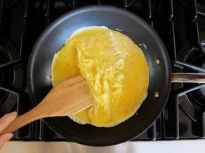 Hand with wooden spatula folding scrambled eggs from outside inward in a nonstick skillet on stovetop.