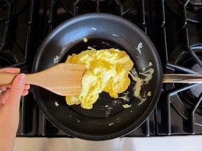 Hand using a wooden spatula to scramble eggs in a nonstick skillet on a stovetop.