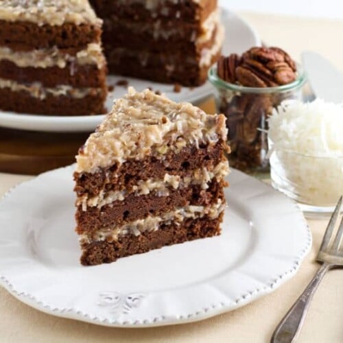 A slice of German Chocolate Cake served on a white plate with coffee grounds, coconut strips in the background