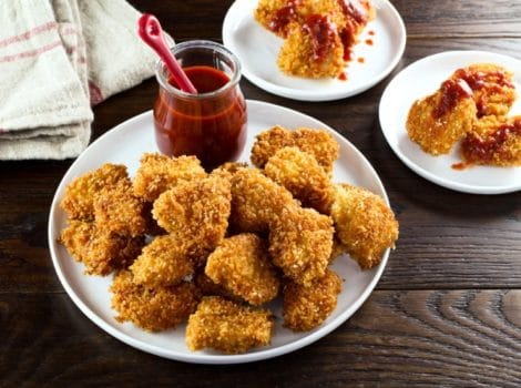 Plate of schnitzel nuggets with red sriracha sauce on wooden table, towel in background.