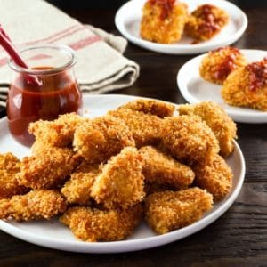 Close up plate of smoky panko schnitzel bites with glass container of sauce, red spoon, on wooden table with sauced schnitzel bites and towel in background.