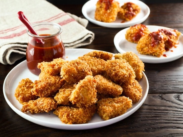 Close up plate of smoky panko schnitzel bites with glass container of sauce, red spoon, on wooden table with sauced schnitzel bites and towel in background.