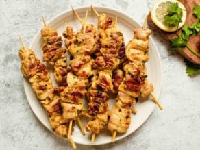 Overhead horizontal shot - plate of Lemony Marinated Chicken Skewers on countertop, lemon and parsley garnish on wooden cutting board beside the plate.