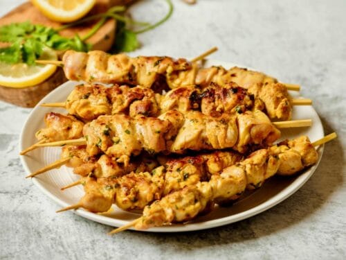 Horizontal shot - plate of Lemony Marinated Chicken Skewers on a marble countertop, wood cutting board with lemon and parsley in background.