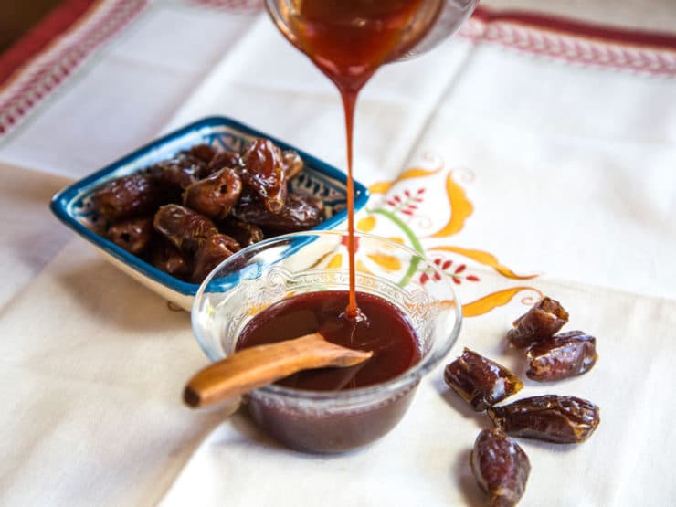 Date Honey Syrup being poured into a glass bowl with wooden spoon, dish of dates in background, scattered dates to the side, on a decorative cloth napkin.