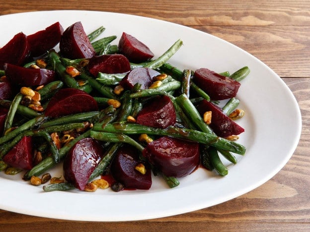 A salad of roasted beets, roasted green beans, and pistachios on a white serving plate atop a wooden table.