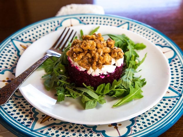 Roasted beet tartare with goat cheese and candied nuts presented on a bed of greens with fork, on a white and decorative Moroccan plate.
