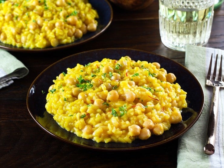 Front angled shot of creamy Saffron Chickpea Risotto on dark blue plate, another plate of risotto, fork, cloth napkin and short decorative wine glass in background.