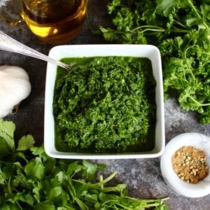 Overhead view of Yemenite Schug sauce in white square bowl with spoon surrounded by fresh herbs, head of garlic, olive oil in glass carafe and spices in white marble bowl on countertop