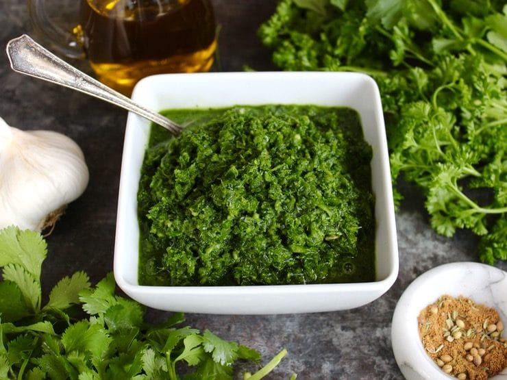 Frontal view of Yemenite Schug sauce in white square bowl with spoon surrounded by fresh herbs, head of garlic, olive oil in glass carafe and spices in white marble bowl on countertop