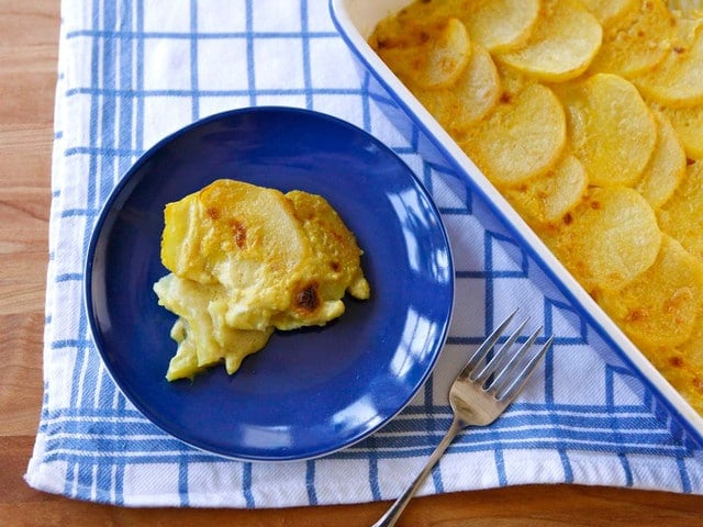 A small serving of scalloped potatoes on a blue plate atop a blue and white checked towel next to a fork and a blue baking dish filled with scalloped potatoes.