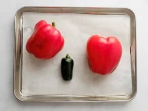Two raw red bell peppers and one raw green chili pepper, on a parchment-lined baking sheet on a marble countertop.