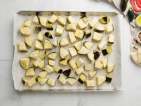 Baking sheet lined with parchment, scattered with cubed raw eggplant pieces.
