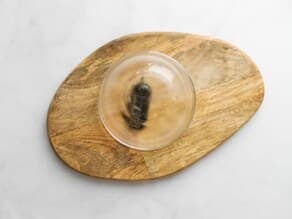Small green chili pepper charred and roasted beneath a steamy small glass bowl on a wooden cutting board, resting on a marble countertop.