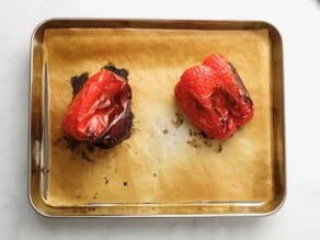 Two red roasted bell peppers, charred and collapsing, on a parchment-lined baking sheet on a marble countertop.