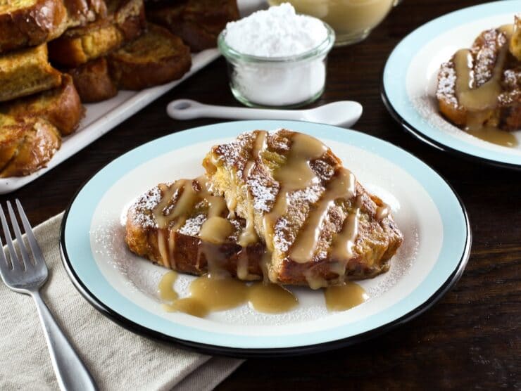 Wider horizontal shot - plate of challah french toast drizzled with Kahlua cream sauce, napkin and platter of french toast in background, powdered sugar with small spoon behind.