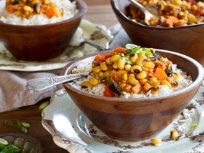 A wooden bowl of vegan hoppin' john black eyed pea stew with spoon on a decorative plate, two more bowls with plates in the background, garnished with green onion.
