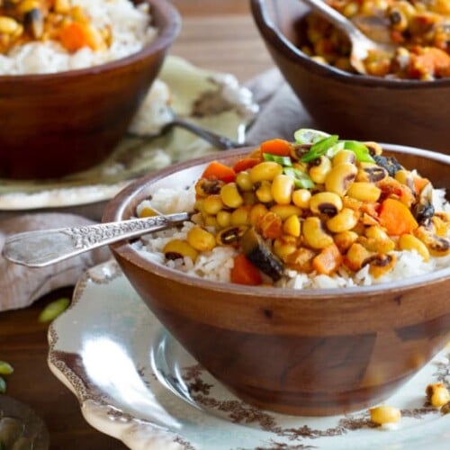 A wooden bowl of vegan hoppin' john black eyed pea stew with spoon on a decorative plate, two more bowls with plates in the background, garnished with green onion.