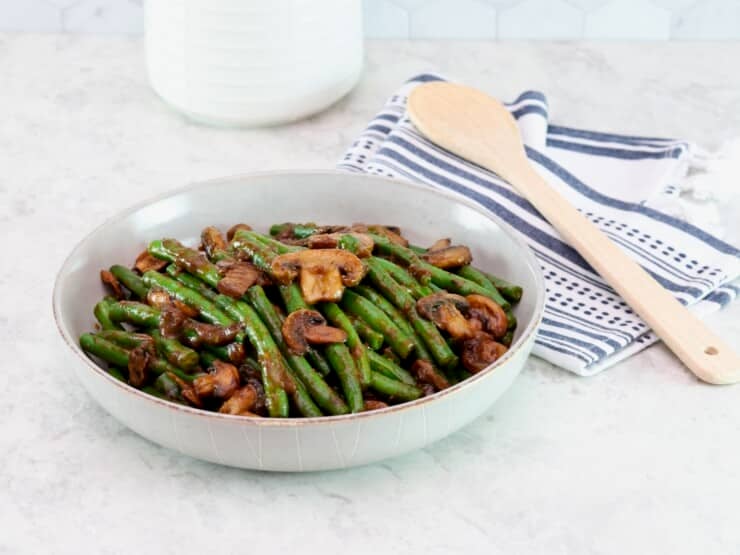 Horizontal frontal shot - large shallow bowl filled with green beans and mushrooms tossed in a rich brown plum sauce. Dish rests on a marble countertop, wooden spoon and kitchen towel beside it. Partial view of white canister in background.