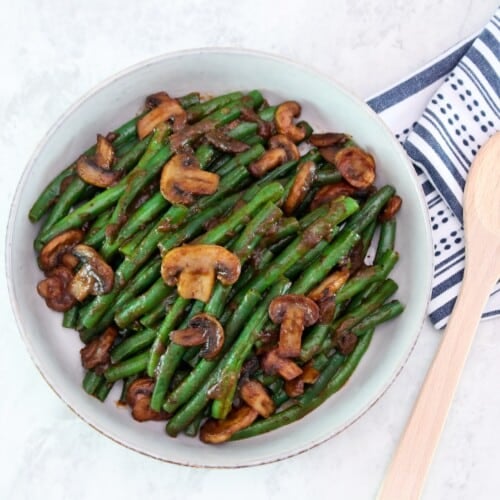 Overhead horizontal shot - large shallow bowl filled with green beans and mushrooms tossed in a rich brown plum sauce. Dish rests on a marble countertop, wooden spoon and kitchen towel beside it.