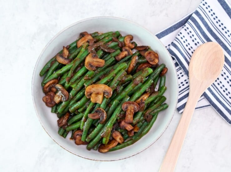 Overhead horizontal shot - large shallow bowl filled with green beans and mushrooms tossed in a rich brown plum sauce. Dish rests on a marble countertop, wooden spoon and kitchen towel beside it.