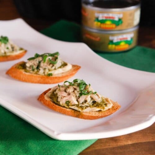 A horizontal cut image of Three Tuna, hummus, and pesto crostini appetizer served on a white plate