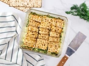 Overhead shot of a spinach matzo mina - savory spinach matzo pie with layers of spinach, feta cheese, and artichokes, baked golden brown. Serving spatula, striped towel, fresh sprig of dill and matzo beside the serving dish.