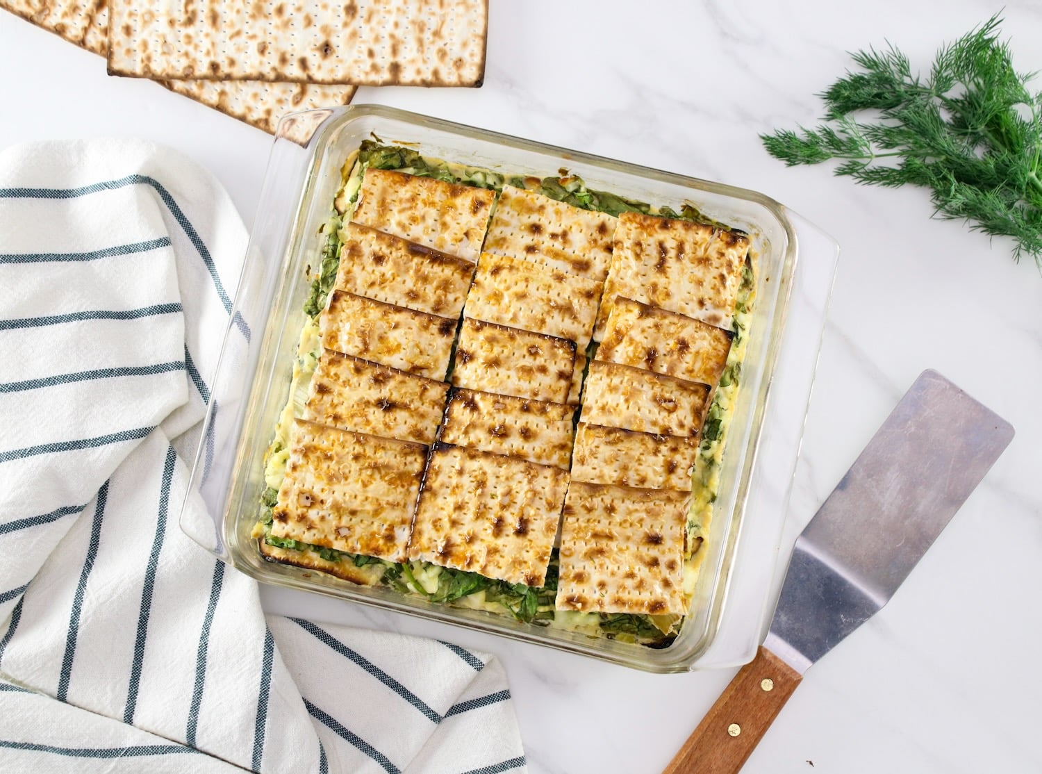 Overhead shot of a spinach matzo mina - savory spinach matzo pie with layers of spinach, feta cheese, and artichokes, baked golden brown. Serving spatula, striped towel, fresh sprig of dill and matzo beside the serving dish.