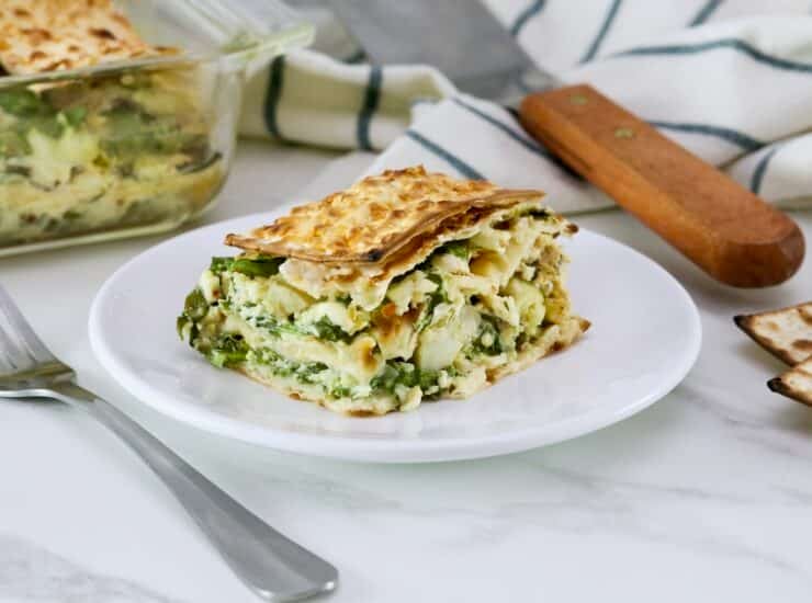 Close up of a slice of spinach matzo pie with layers of spinach, feta cheese, and artichokes, baked to perfection and served on a white plate, fork and towel beside the plate. Spinach matzo mina pie in background.