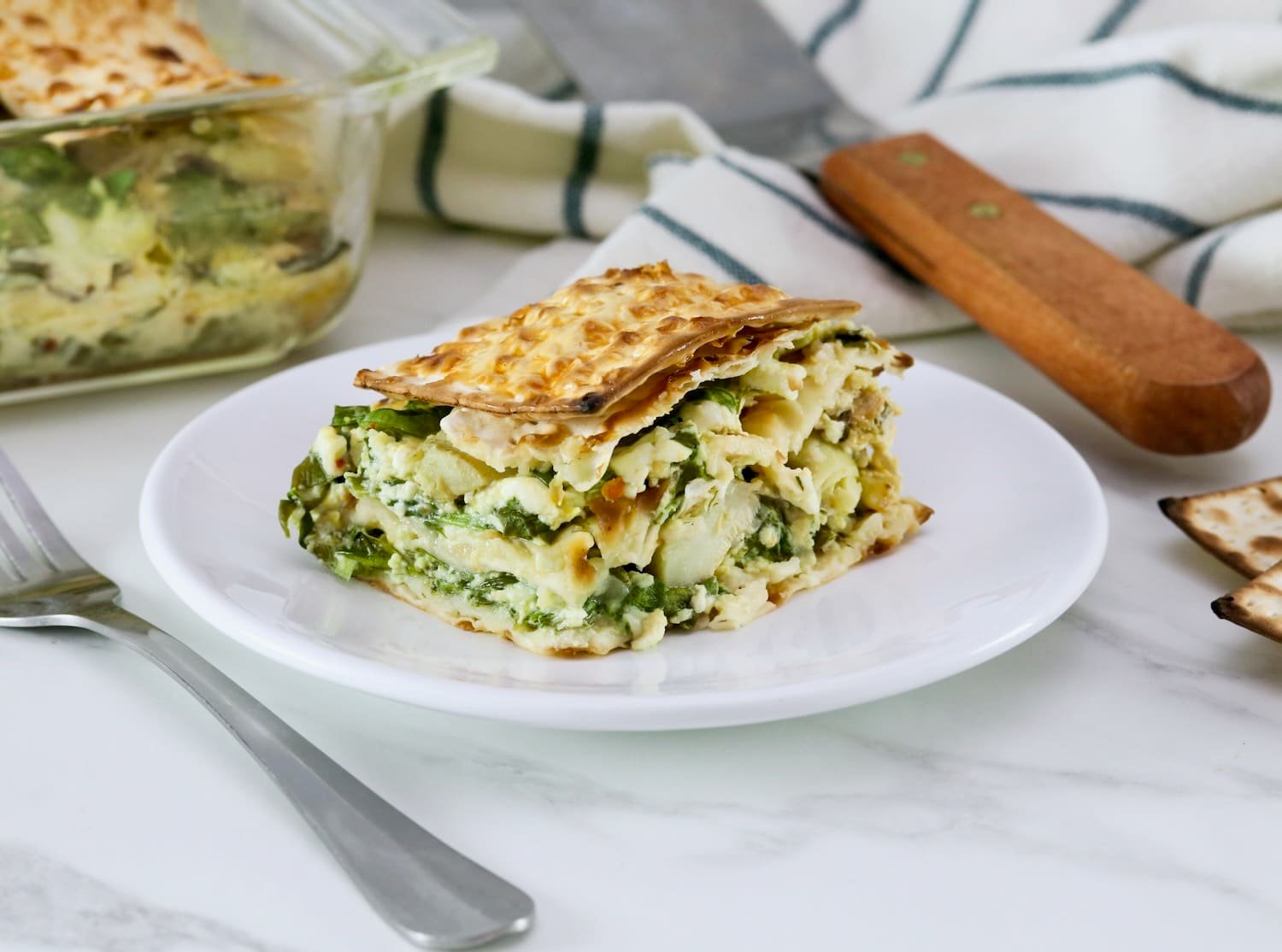 Close up of a slice of spinach matzo pie with layers of spinach, feta cheese, and artichokes, baked to perfection and served on a white plate, fork and towel beside the plate. Spinach matzo mina pie in background.