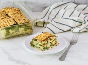 A savory spinach matzo pie in square casserole dish, a slice of spinach matzo pie with layers of spinach, feta cheese, and artichokes, baked to perfection and served on a white plate, fork and towel beside the plate.