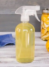 Spray bottle filled with all-natural all-purpose cleanser, jar of steeping peels and whole lemon in background with blue towel.