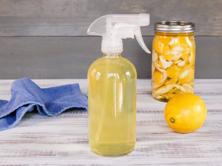 Spray bottle filled with all-natural all-purpose cleanser, jar of steeping peels and whole lemon in background with blue towel.