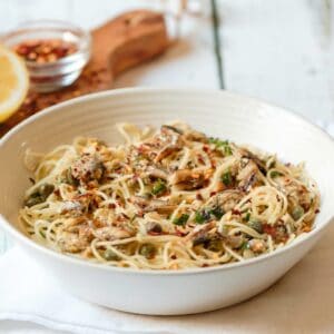 Front view horizontal shot - white bowl of sardine pasta capellini garnished with chili flakes and fresh parsley, resting on a white cloth napkin, wooden cutting board in background with lemon half and chili flakes, on white wood table.