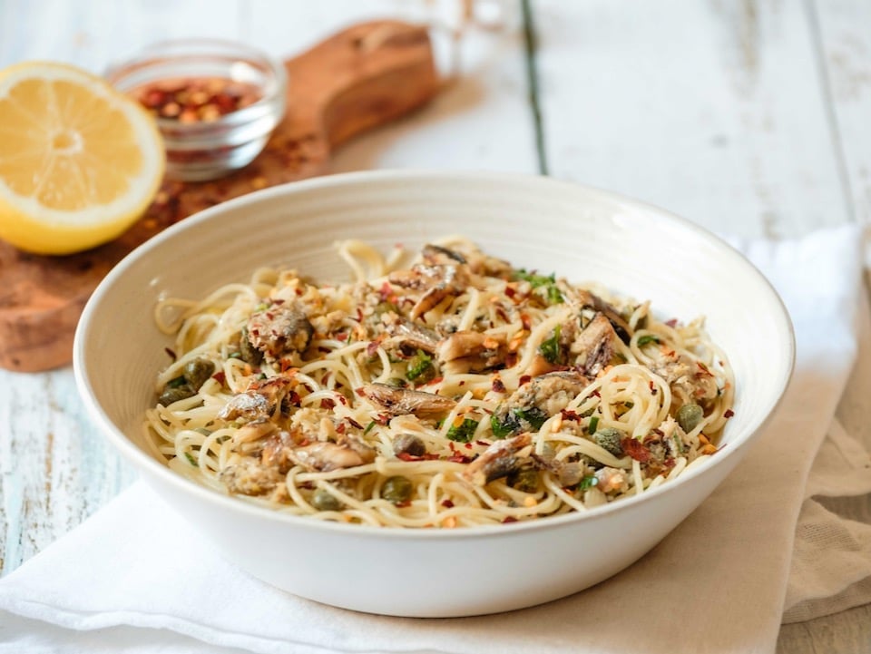 Front view horizontal shot - white bowl of sardine pasta capellini garnished with chili flakes and fresh parsley, resting on a white cloth napkin, wooden cutting board in background with lemon half and chili flakes, on white wood table.