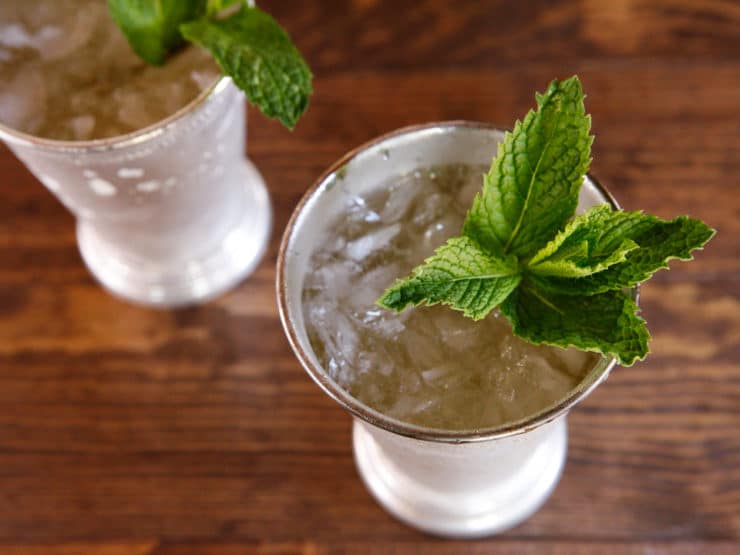 Two glasses of Mint Julep, filled with ice and garnished with fresh mint leaves, placed on a rustic wooden table